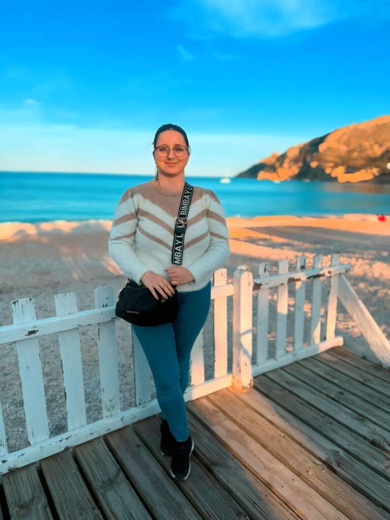 ristina disfrutando de un día soleado en la playa, mostrando su estilo relajado con un suéter blanco y gafas de sol, mientras posa frente al mar.