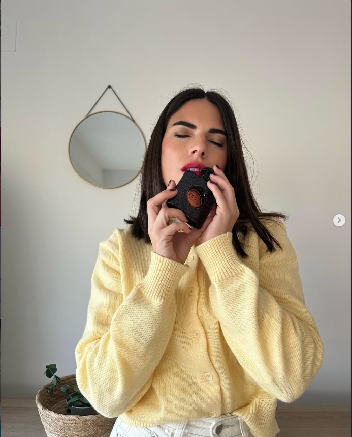 Laura Toledo con suéter amarillo sosteniendo un producto, con expresión relajada y un fondo minimalista.