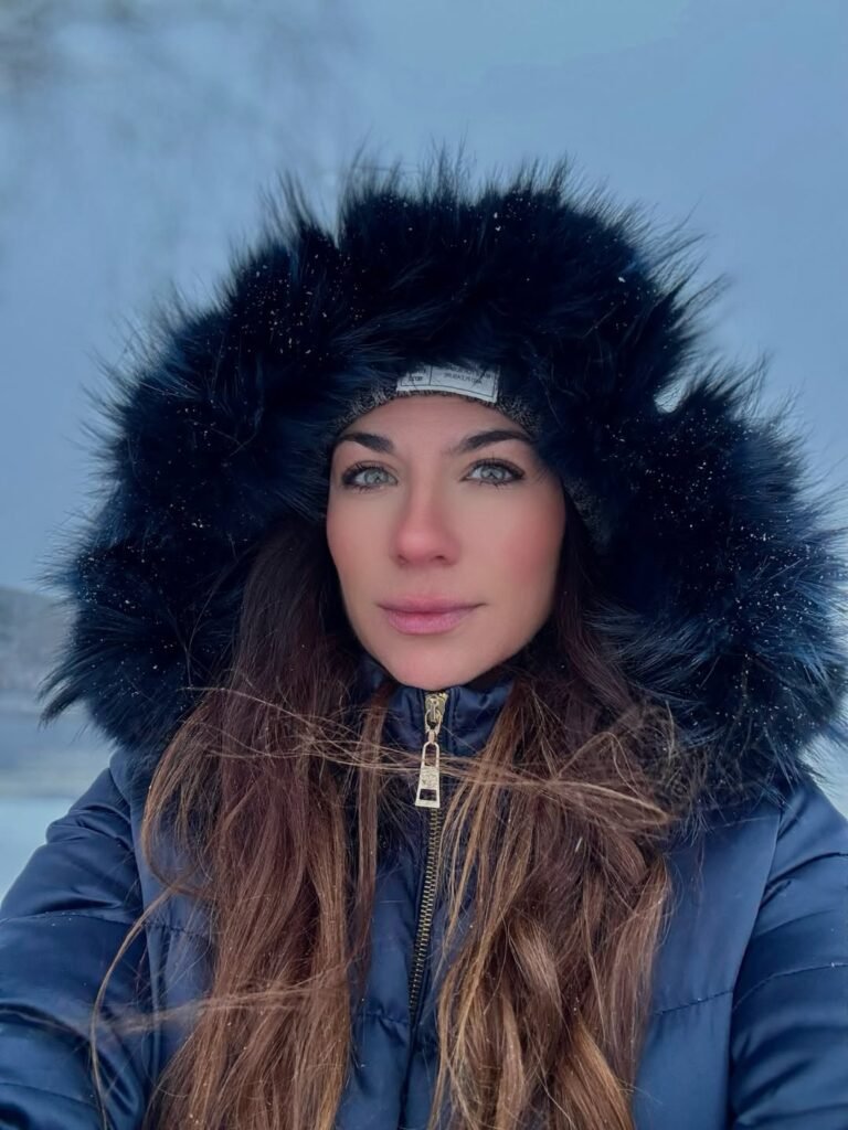 Cristina Jiménez sonríe frente a un edificio moderno, vistiendo un top negro brillante y pantalones oscuros, capturando un momento lleno de energía en la ciudad.