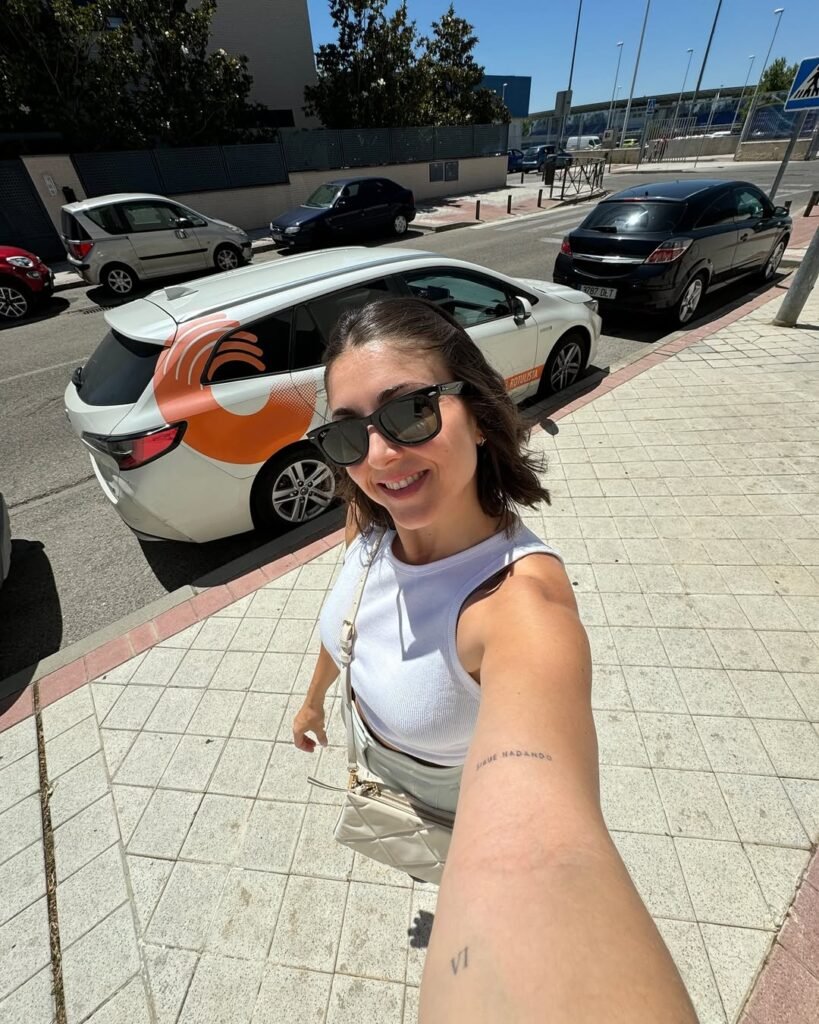 Alba Garcia Diez posando en la calle, usando gafas de sol y una camiseta blanca, frente a un coche con logotipo en el fondo.