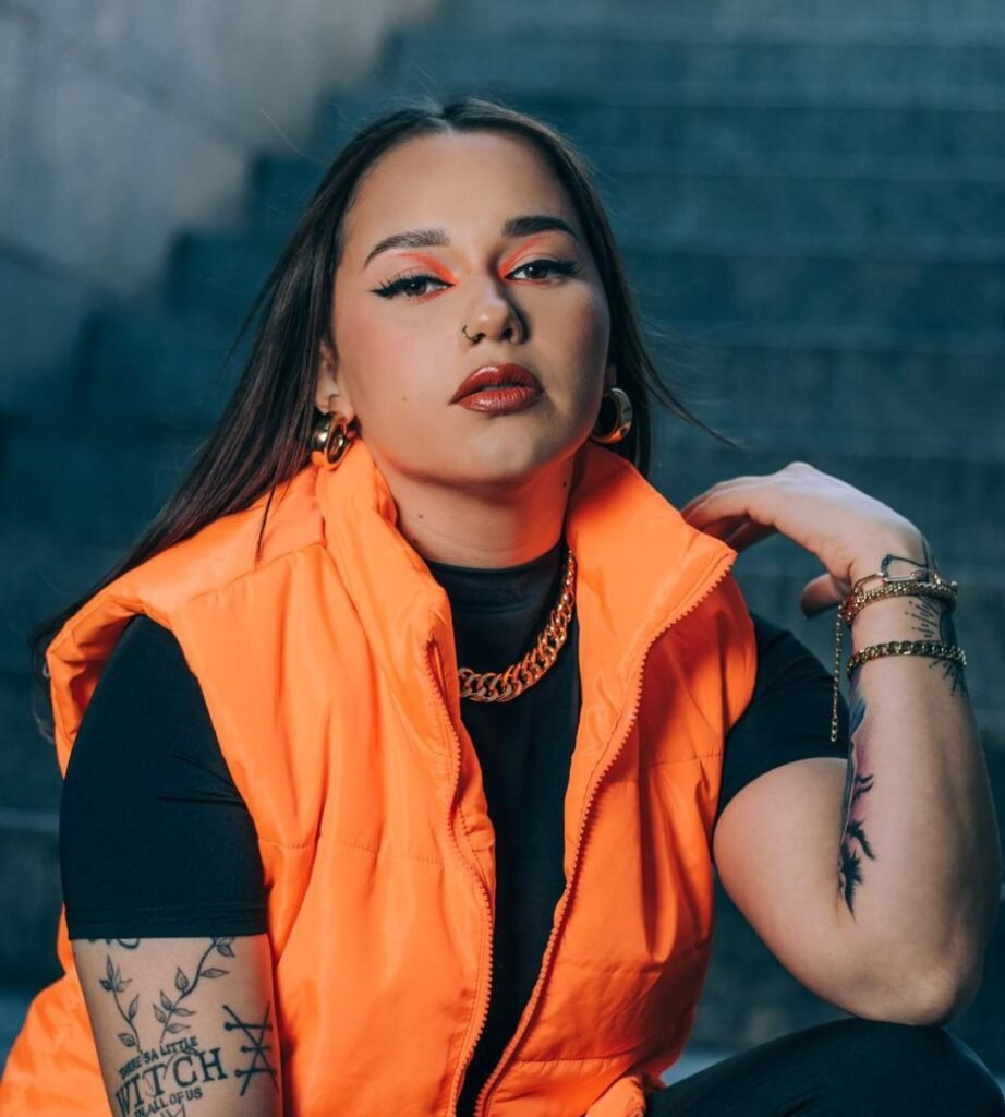 Barbara Elena posando con chaleco naranja y maquillaje llamativo, mostrando su estilo único y actitud audaz.