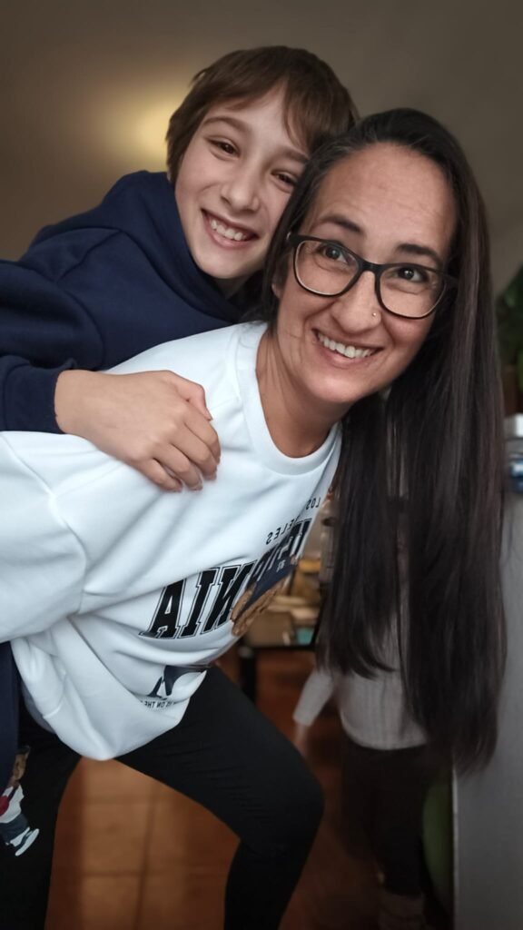 Cristina Asensio Aranda sonriendo mientras su hijo pequeño está sobre su espalda, ambos luciendo ropa cómoda y casual.