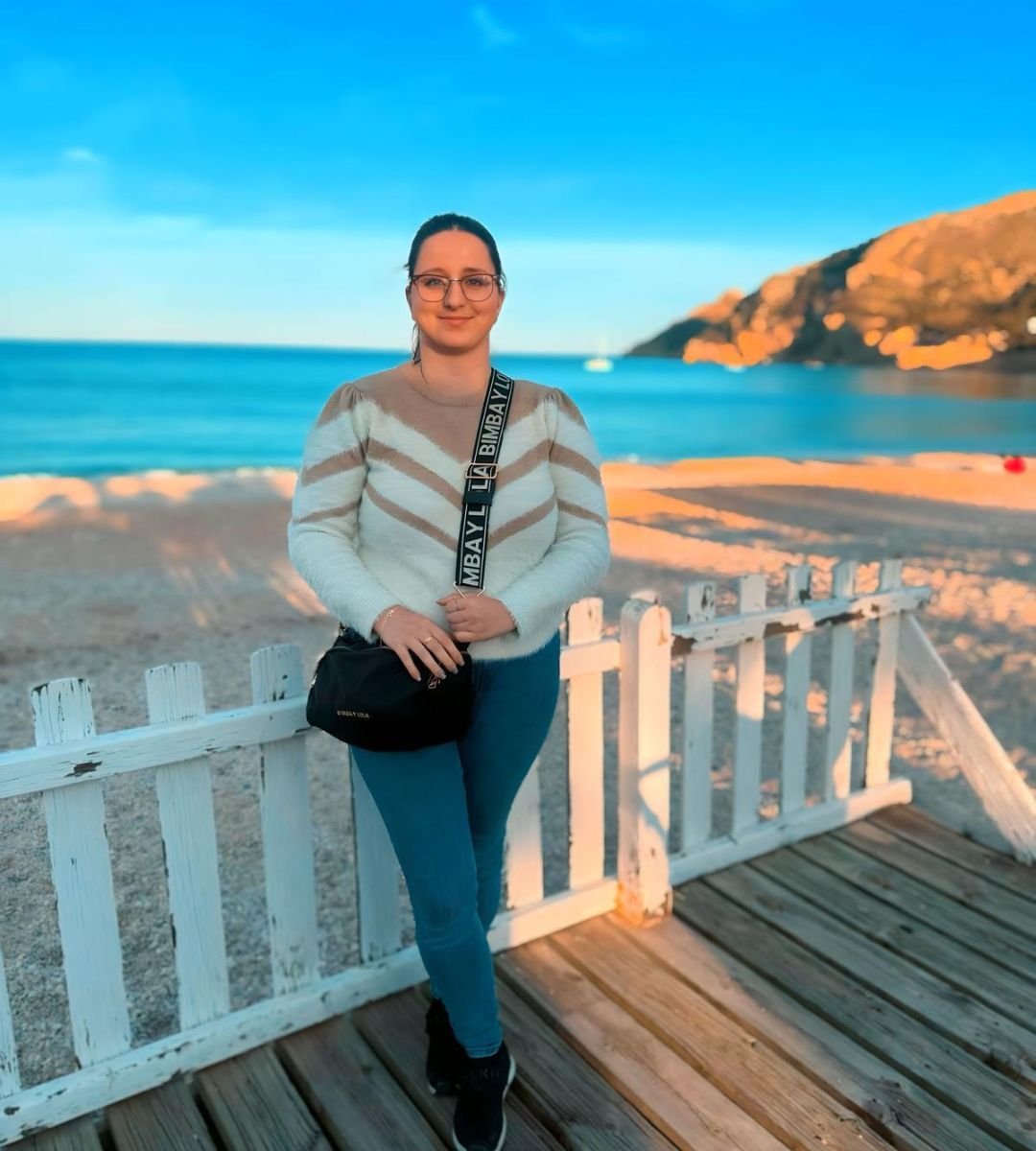 Cristina Pérez, creadora de contenido UGC, posando con suéter blanco, jeans azules y bolso negro en la playa, disfrutando de un paisaje al atardecer.