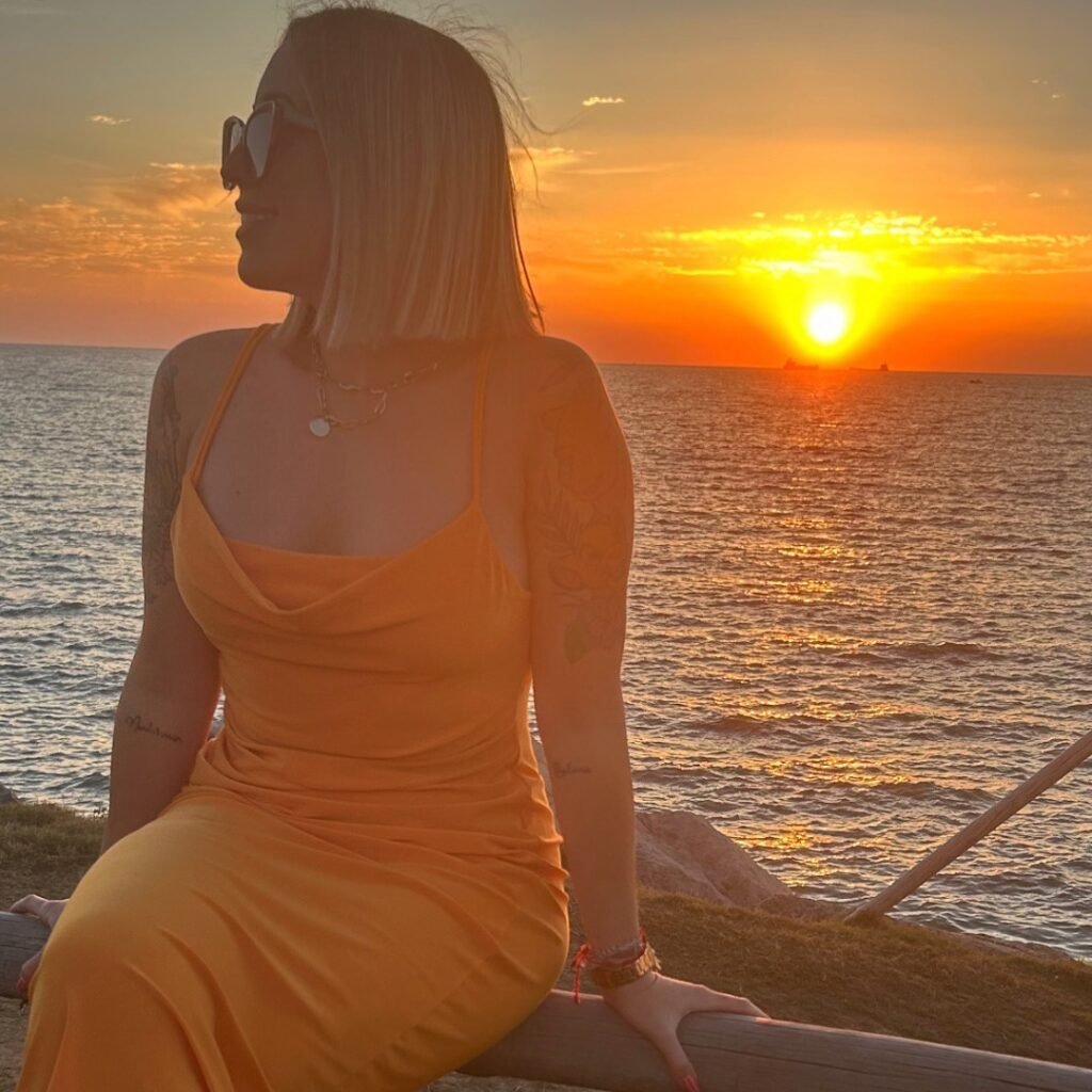 Laura Domínguez González sentada frente a un atardecer sobre el mar, usando gafas de sol y un vestido amarillo, con un estilo relajado y elegante.