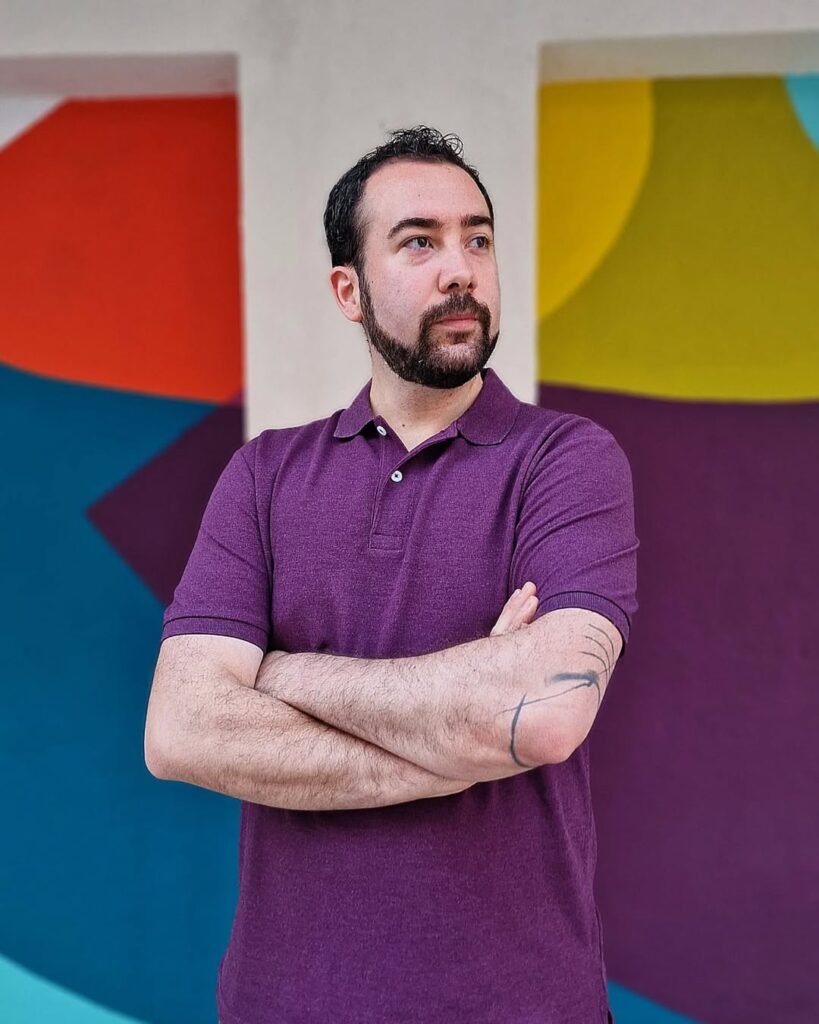 Miguel Tarjuelo posando con los brazos cruzados frente a un mural colorido, con una camiseta púrpura.