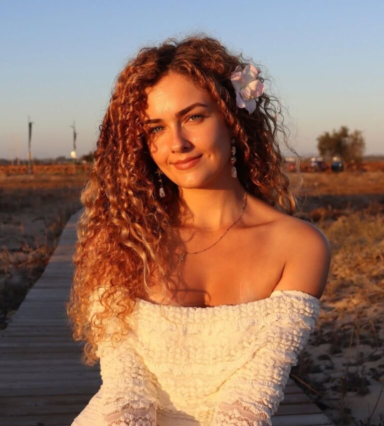 Cristina Ruiz Marin sonríe al atardecer con flores en el cabello, mientras posa frente a un hermoso paisaje en la playa. Foto perfecta para una sensación natural y luminosa.