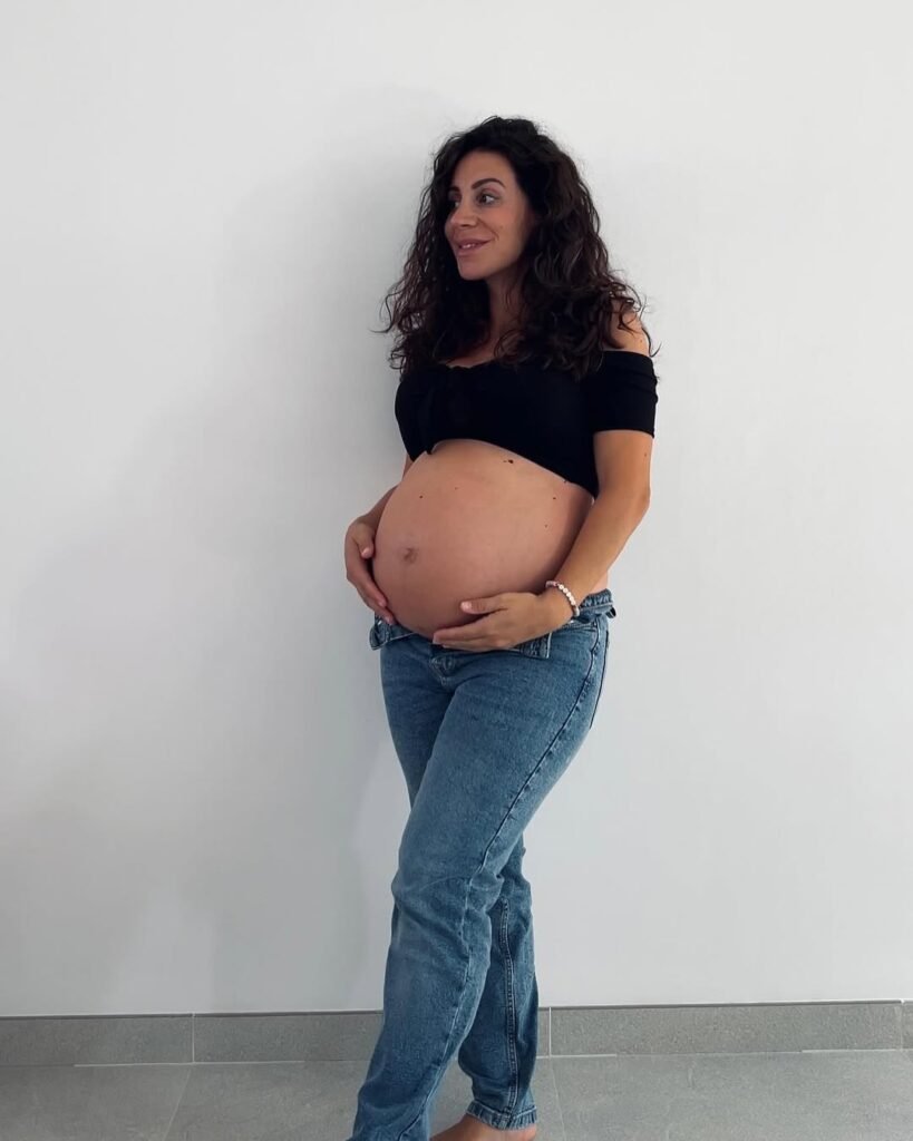 Cinzia Ledonne posando embarazada con jeans, tocando su barriga con fondo blanco.