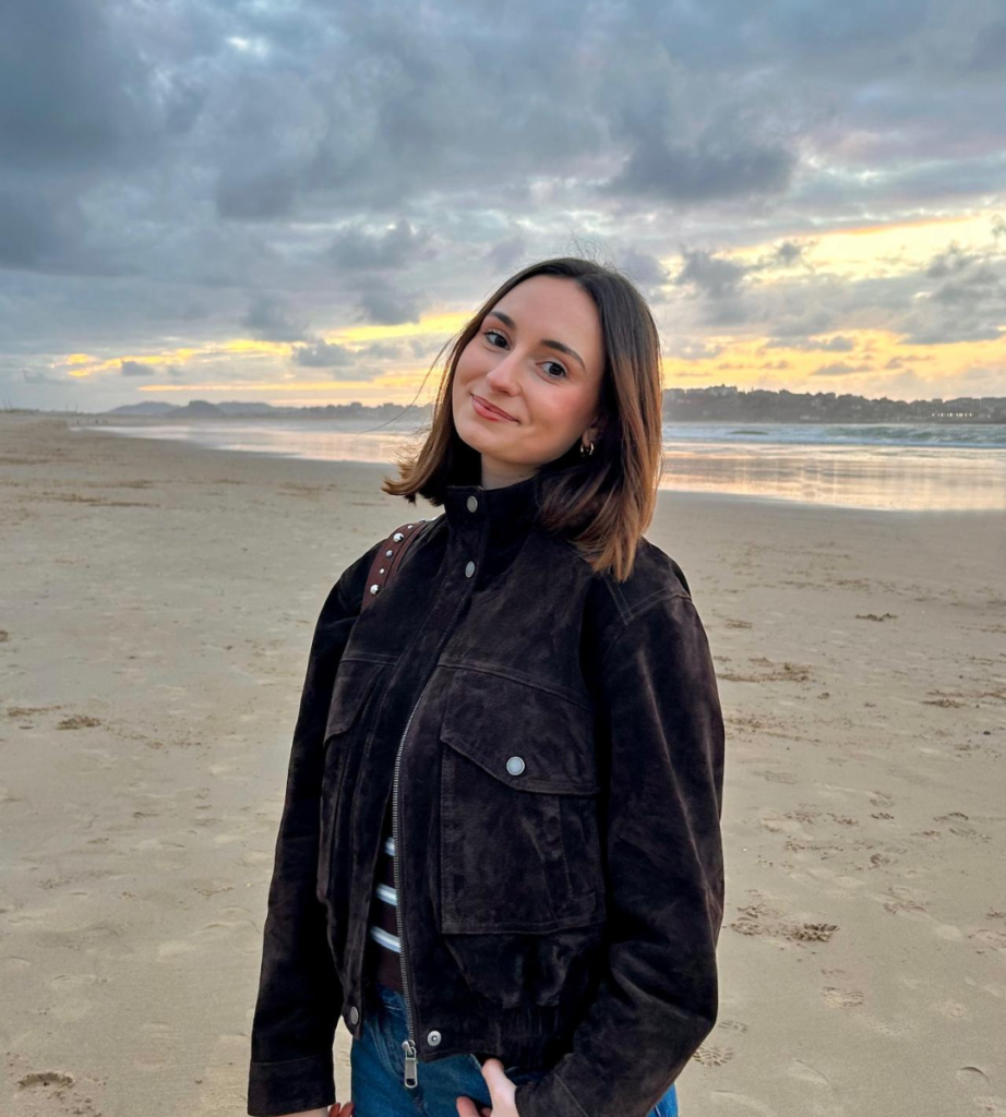 Aida, creadora de contenido, posando en la playa con una chaqueta marrón, representando su estilo auténtico y relajado.