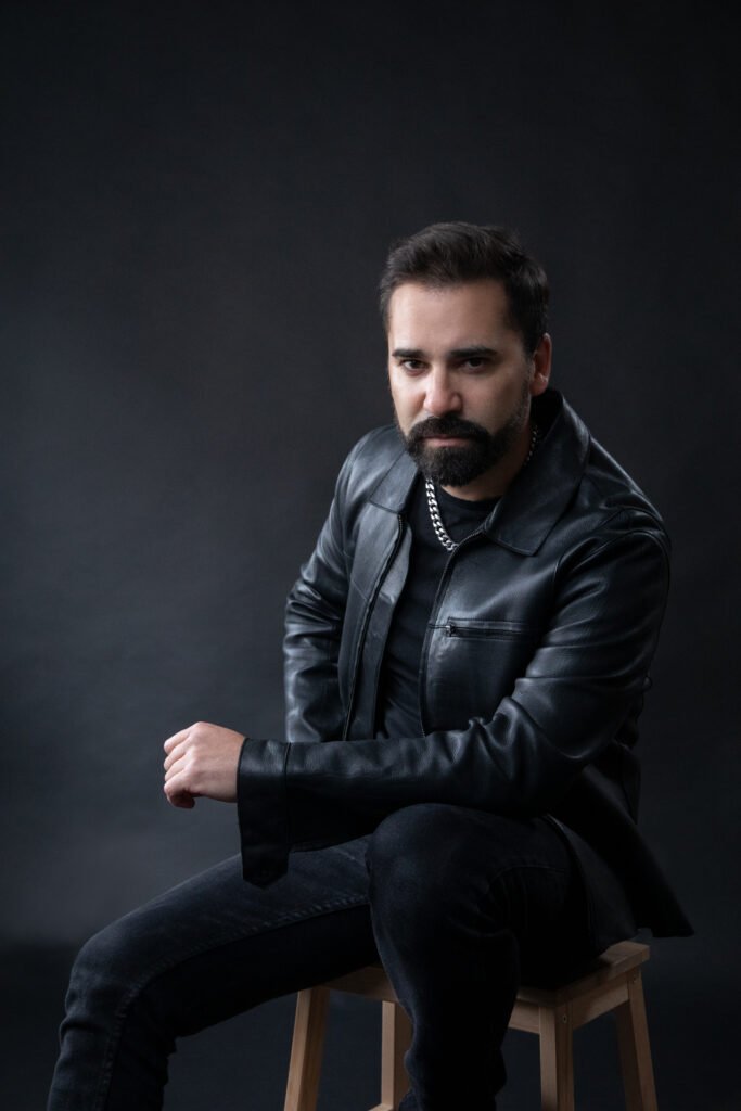 Hombre adulto con barba y chaqueta de cuero negra sentado sobre taburete en fondo oscuro