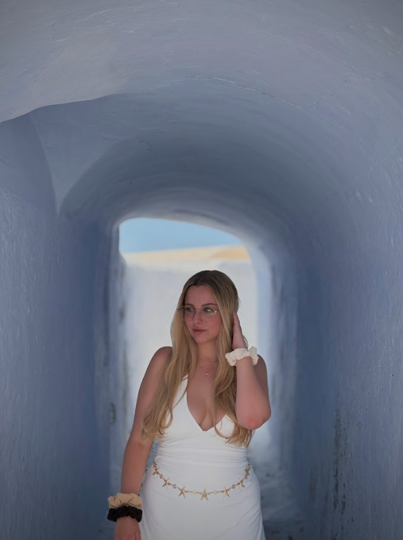Mujer rubia con vestido blanco caminando por un túnel pintado de blanco, con luz natural al fondo.