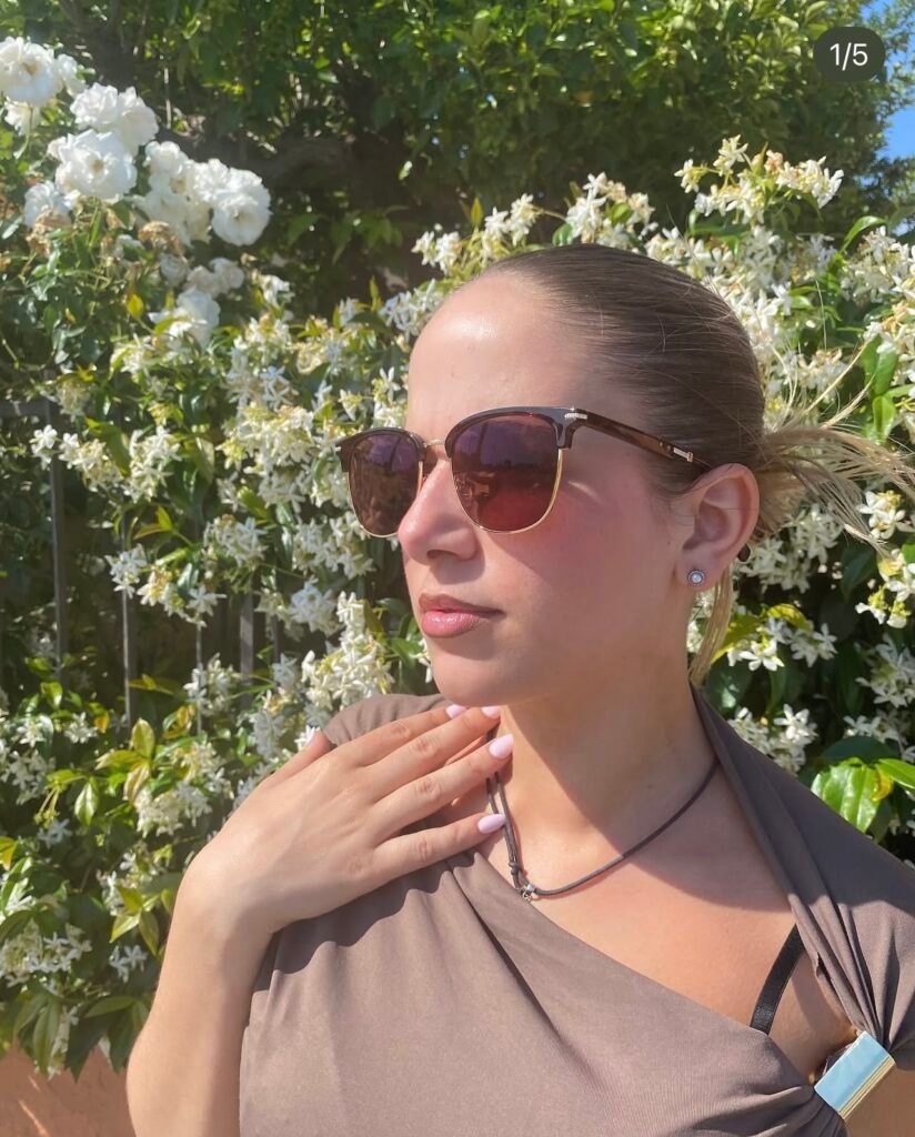 Natalia con gafas de sol posando frente a arbustos con flores blancas al aire libre.