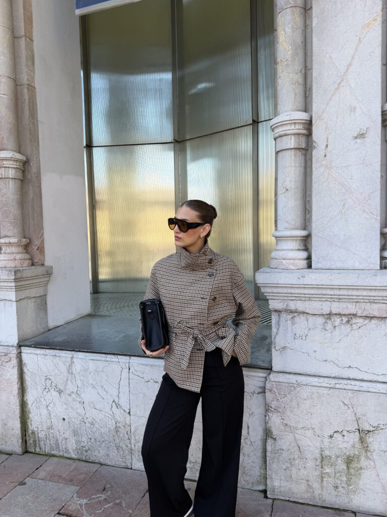 Mujer con gafas de sol y abrigo a cuadros, posando frente a una pared de mármol.