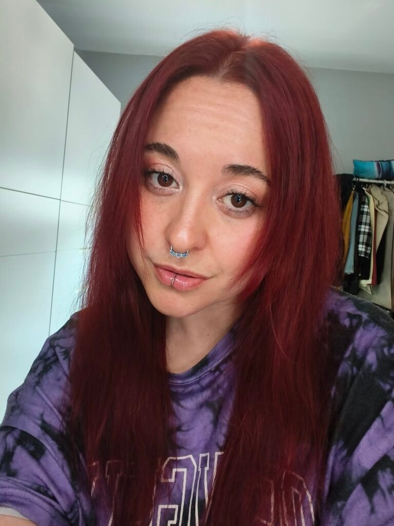 Chica joven con cabello rojo liso, piercing en la nariz y camiseta morada posando en una selfie.