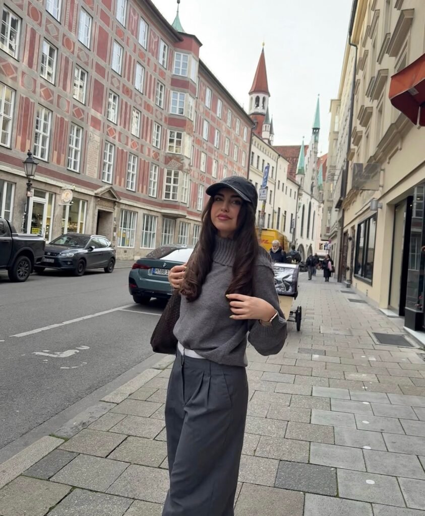 Mujer con suéter gris y gorra negra posando en la calle de una ciudad europea con arquitectura histórica.