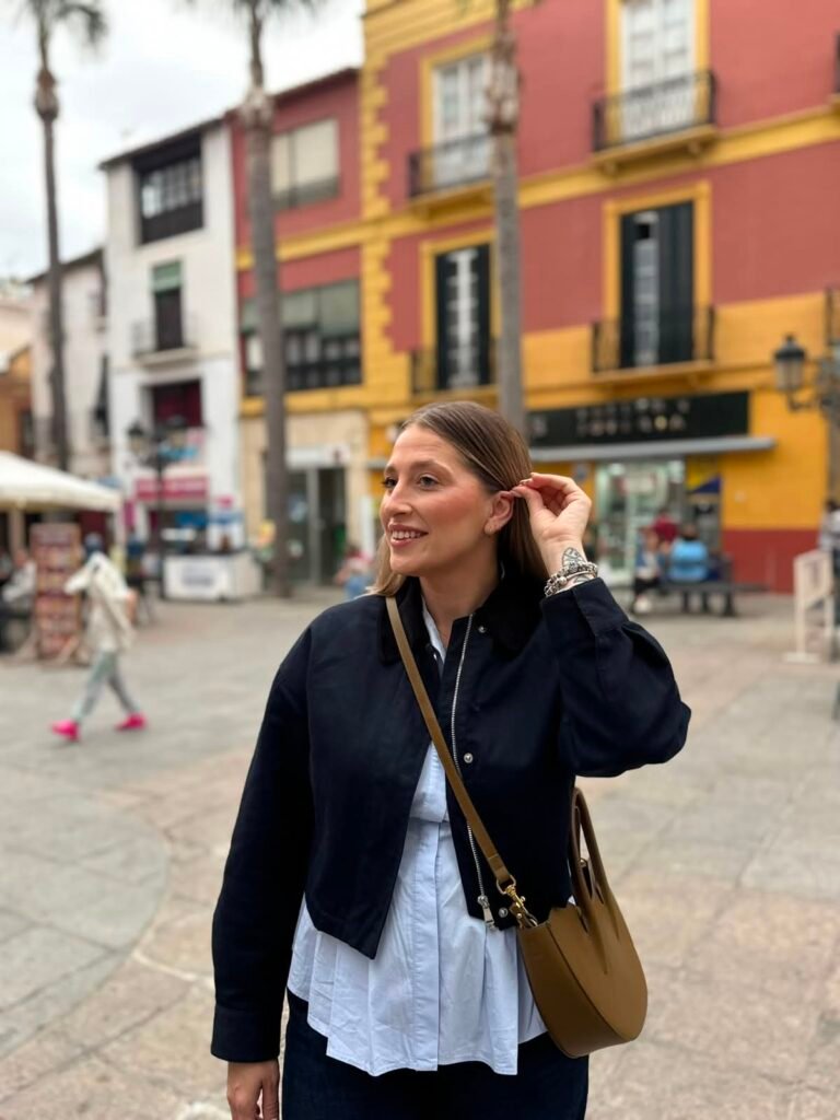 Mujer en la calle sonriendo y ajustando su cabello, con un bolso y un fondo colorido de edificios.
