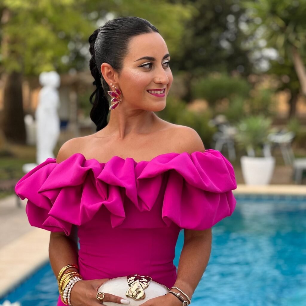 Laura Lobato posando junto a una piscina con vestido fucsia con volantes y accesorios dorados, mostrando estilo y elegancia.