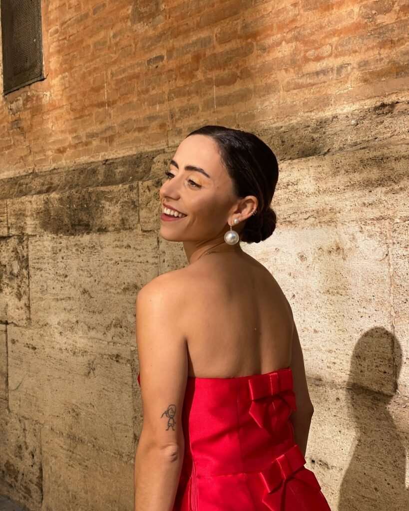 Eli sonriente de perfil con vestido rojo elegante y moño, mostrando un maquillaje natural y pendientes llamativos, fotografía al aire libre.