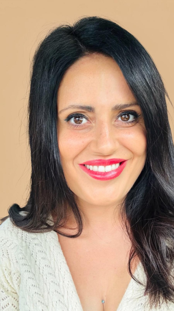 Mujer con cabello oscuro y largo, sonriendo frente a fondo beige, mostrando maquillaje natural y labios rojos.