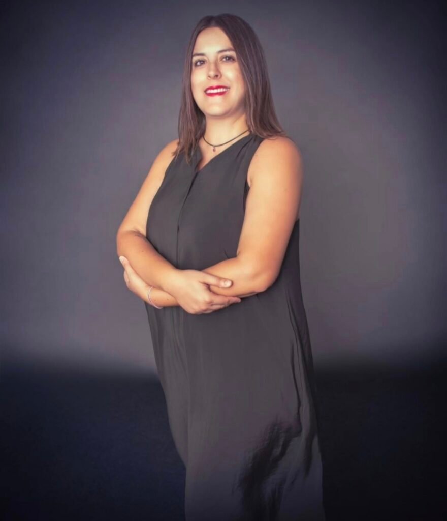 Andrea Renau Vázquez posando frente a un fondo gris con los brazos cruzados, mirada segura y sonrisa profesional.