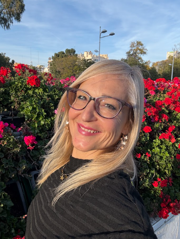 Mujer rubia sonriente con gafas frente a flores rojas al aire libre