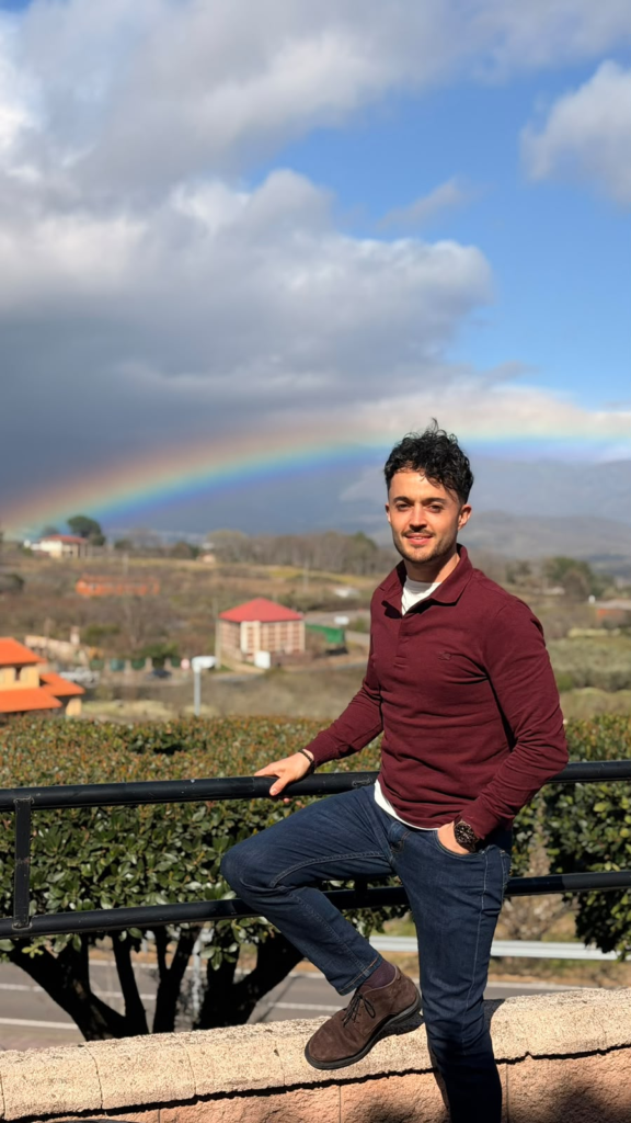 Julio Alcón García posando al aire libre con un paisaje natural de fondo y un arcoíris visible en el horizonte. Luce un outfit casual elegante compuesto por jersey granate, vaqueros oscuros y zapatos de piel.