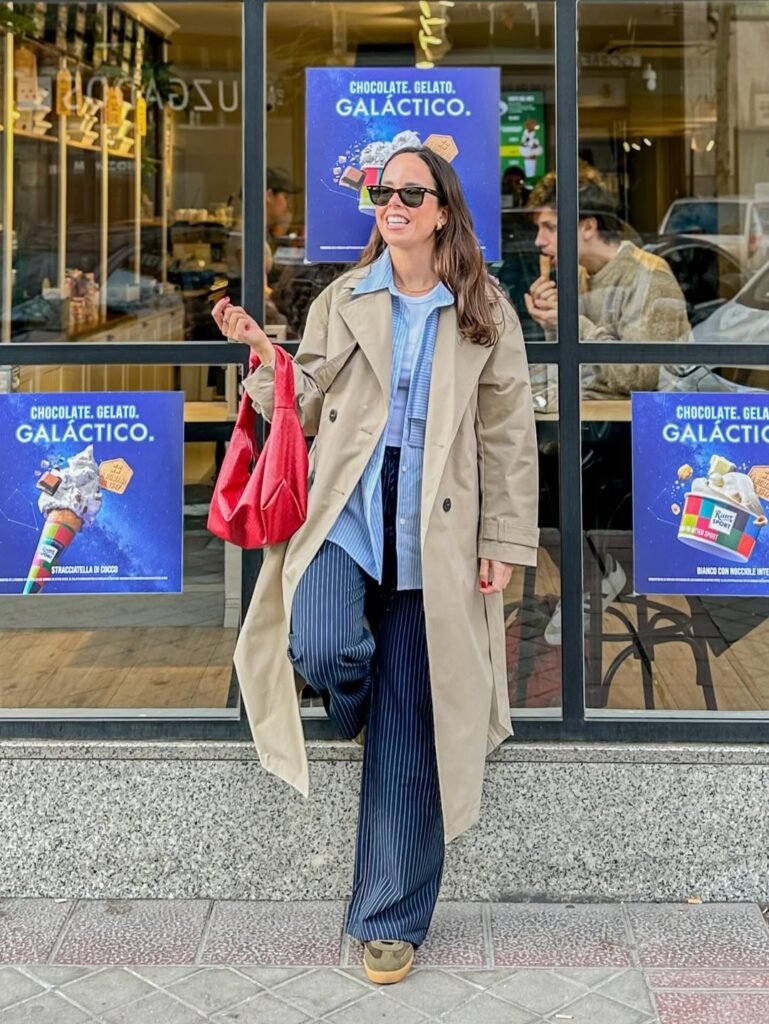María Huidobro, creadora de contenido UGC lifestyle, posando frente a una cafetería con un estilismo urbano y elegante compuesto por gabardina beige, conjunto azul de rayas y bolso rojo, transmitiendo una imagen moderna y sofisticada.
