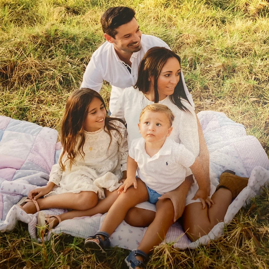 Familia feliz sentada sobre una manta en el césped, con padre, madre, hija y hijo pequeño sonriendo y disfrutando del momento al aire libre.