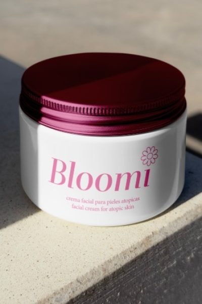 Crema facial Bloomi para pieles atópicas, envase blanco con detalles rosa, sobre fondo claro.