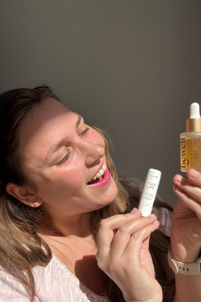 Casandra Benassar sosteniendo un serum y un bálsamo labial, sonriente y mirando hacia un lado, luz natural sobre su rostro.