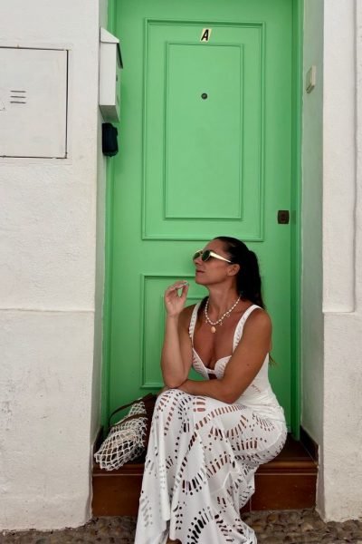 Agencia de marketing digital Brilla Influencers y Comunicación, mujer con vestido blanco de encaje, sentada frente a puerta verde, con gafas de sol y bolso de mano.