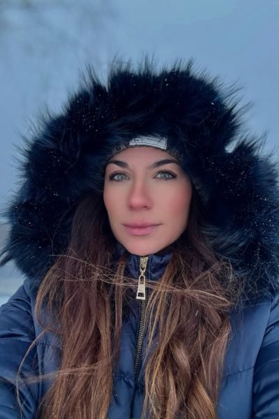 Cristina Jiménez sonríe frente a un edificio moderno, vistiendo un top negro brillante y pantalones oscuros, capturando un momento lleno de energía en la ciudad.