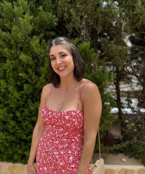 Alba García Diez posando con un vestido rojo con estampado floral frente a un fondo de árboles, sonriendo.