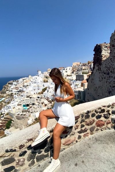 Alicia García sentada en el borde de un muro en Santorini, con un conjunto blanco y tenis blancos.