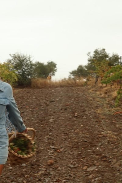 Alicia García caminando por un campo con una canasta y chaqueta de mezclilla.