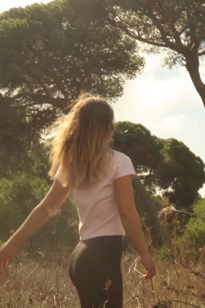 Alicia García caminando entre árboles bajo la luz del sol, con una camiseta rosa y pantalones negros.