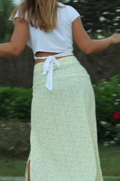 Alicia García de espaldas en el parque, usando un top blanco y falda larga con detalles florales.