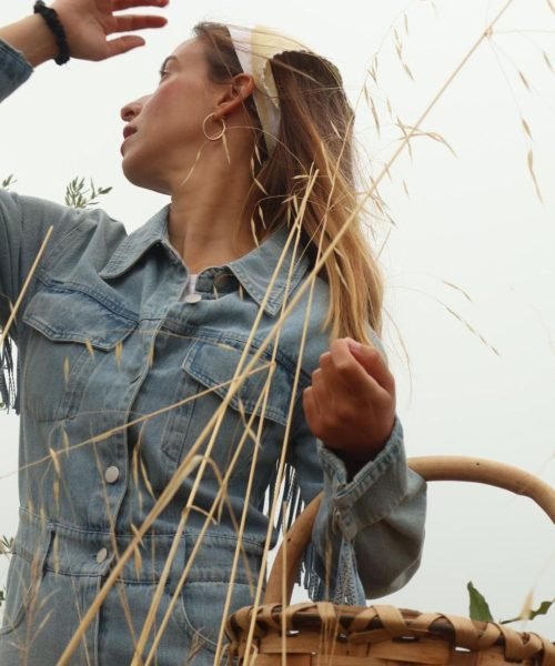 licia García en un campo, mirando hacia el horizonte mientras sostiene una canasta, con una chaqueta de mezclilla y diadema.