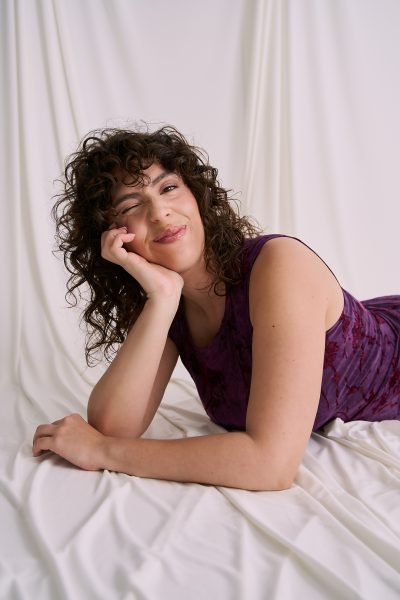 Ana Carpio recostada sobre un fondo de tela blanca con vestido morado, guiñando un ojo y sonriendo de forma natural durante una sesión editorial.