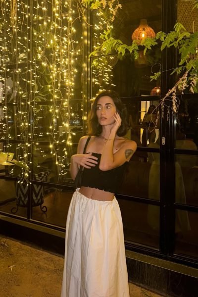 Ana Flores Campos posando en un espacio con iluminación cálida y luces decorativas, luciendo un conjunto elegante de top negro y falda blanca para una sesión de moda lifestyle nocturna.