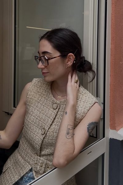 Ana Flores Campos con gafas y chaleco de tweed beige en una sesión de moda casual elegante junto a una ventana.
