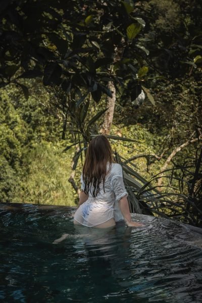 Arantxa Styles en la piscina con una camisa blanca, mirando hacia atrás, rodeada de naturaleza.