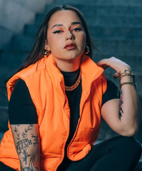 Bárbara Elena con chaleco naranja, mostrando una actitud fuerte y moderna en un fondo oscuro.