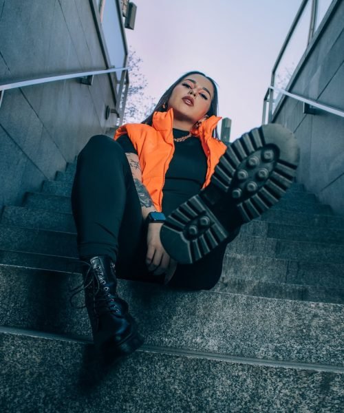 Bárbara Elena posando en las escaleras con chaleco naranja, botas negras y actitud desafiante.