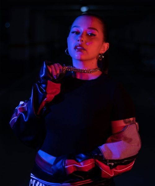 Bárbara Elena con chaqueta de cuero, maquillaje llamativo y luz de neón, mostrando actitud y estilo en un ambiente oscuro.