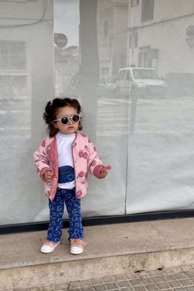 Niña pequeña con gafas de sol, posando en la calle con chaqueta rosa.