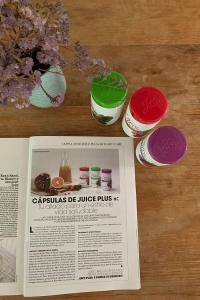 UGC de Cristina Agudo Plaza con cápsulas Juice Plus sobre una mesa de madera, acompañadas de un florero y una revista abierta.