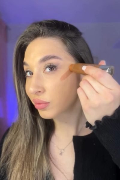 UGC de Cristina Agudo Plaza aplicando maquillaje en el rostro con la mano, con fondo interior y luz cálida.