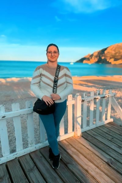 ristina disfrutando de un día soleado en la playa, mostrando su estilo relajado con un suéter blanco y gafas de sol, mientras posa frente al mar.