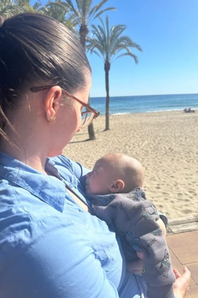Cristina abrazando a su bebé en la playa, disfrutando de un día soleado junto a su hijo, con un fondo de palmeras y el mar de fondo.