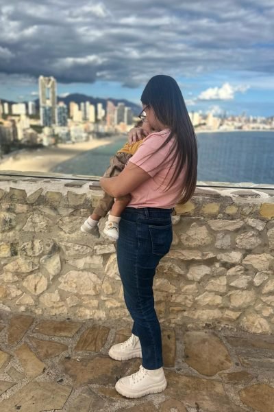 Cristina sosteniendo a su bebé mientras observa el horizonte desde una altura con la ciudad de Benidorm de fondo.